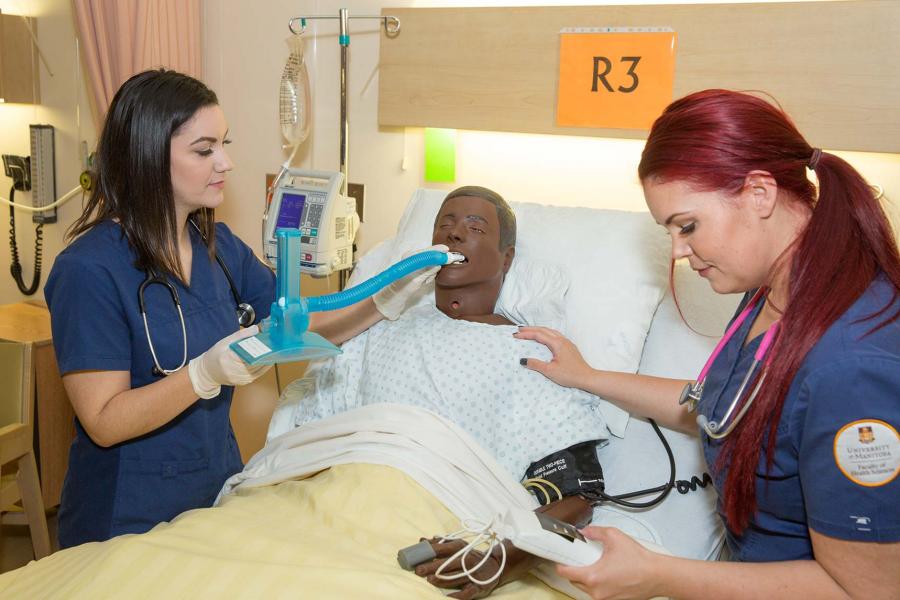 Students wearing scrubs in a hospital room work on a mannequin wearing a hospital gown in a hospital bed.