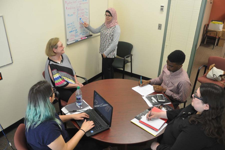Four people sitting around a circular table in a small room taking notes while an instructor wearing a hijab stands pointing at a white board.