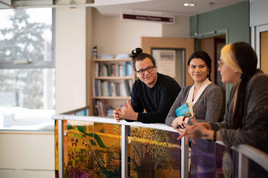 Three Indigenous students leaning against a balcony on the second floor of Migizi Agamik. The glass below the balcony railing depicts colourful nature scenes.