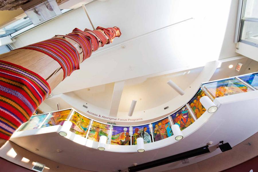 Perspective looking up from the centre of the Migizi Agamic Foyer, along the length of a tall wooden post wrapped in a Métis sash. Along the second floor balcony are a series of colourful paintings and a sign reading "Access and Aboriginal Focus Programs".