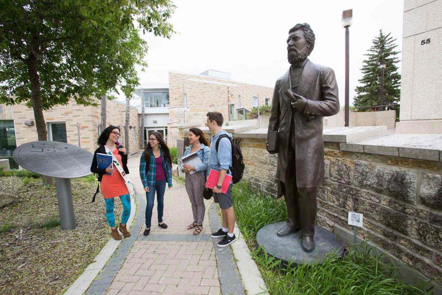 Four Indigenous students standing on a brick walking path in front of Migizii Agamik – Bald Eagle Lodge. To their right is a bronze statue of Louis Riel.