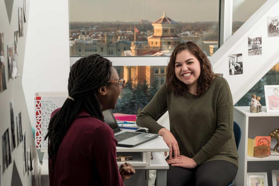 Two students sit laughing in a Pembina Hall dorm room. The room has white walls and furnishings with large windows overlooking the Fort Garry campus.