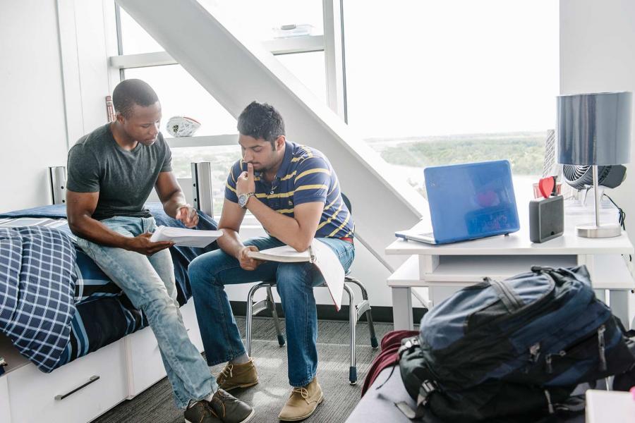 Two students studying in a Pembina Hall dorm room. One is sitting on a bed and the other is sitting on a desk chair. The furnishings are white and modern, and large windows overlook the Red River. 