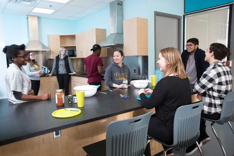 Eight students gathering in a large shared dorm kitchen. Some sit at tall chairs while others prepare food. 