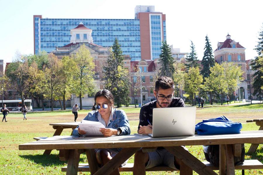 Two students sitting with laptop and papers studying at an outdoor picnic table on a lawn. The UM Admin building and Pembina Hall Residence are behind them. 