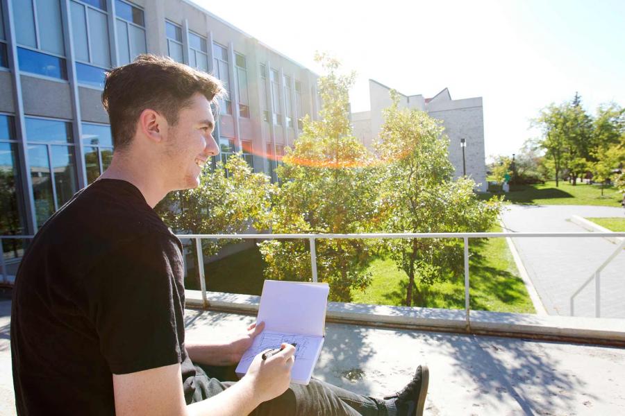 A student with short hair and a black t-shirt sits outside holding a pencil and sketching notebook while smiling and looking out of frame.  