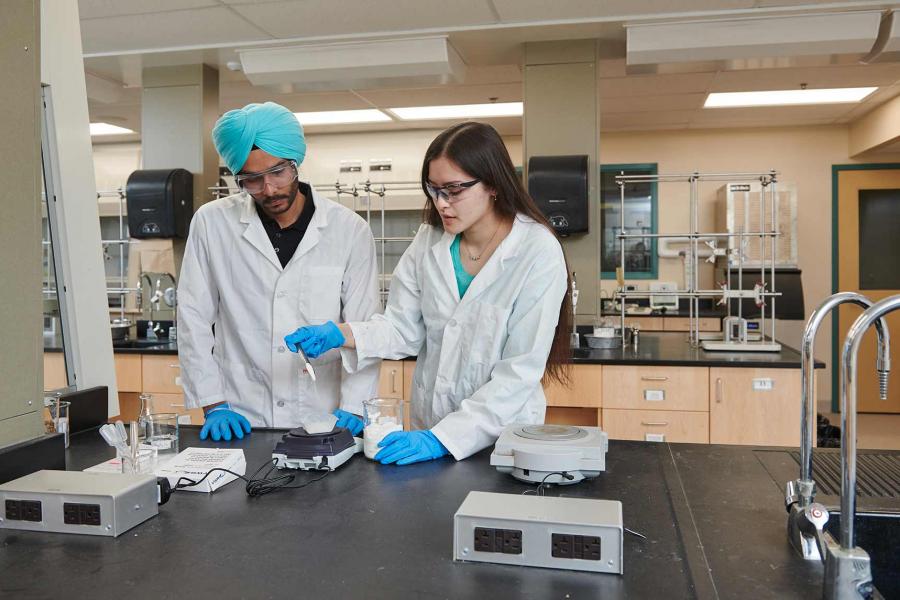 Two students wearing protective gloves, glasses and lab coats stand in a scientific lab. One student, wearing a turban, watches as the other transfers a chemical from one container to another.