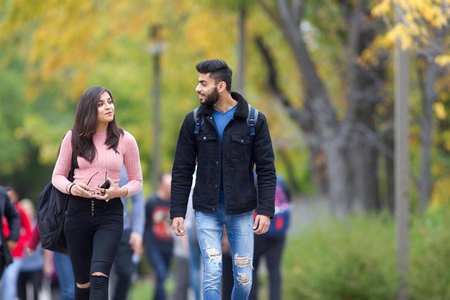 Two students holding bags look at each other in conversation while walking outside toward the camera. The student on the left has long brown hair, a pink shirt and black jeans. The student on the right has dark hair and a beard and is wearing a black casual jacket and blue jeans.  