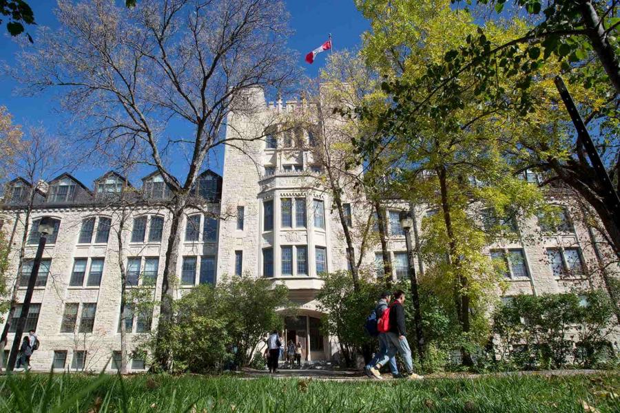 A large, stately limestone brick building with many windows and a Canadian flag, surrounded by trees. 