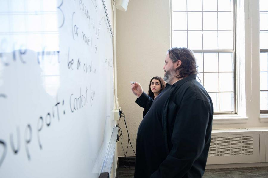 A bearded instructor writes on a whiteboard while a student looks on.