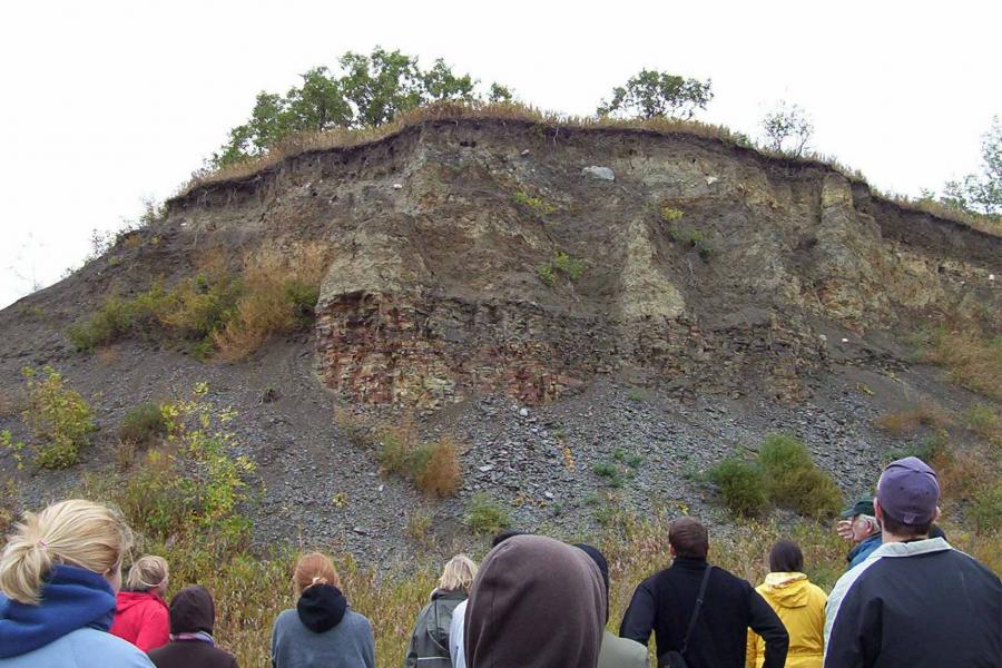 A group of students on a study trip look at layers of sediment on the side of a cliff.