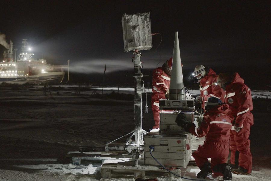 Four arctic researchers analyze technical equipment at night in the snow, with a large ship in the background.