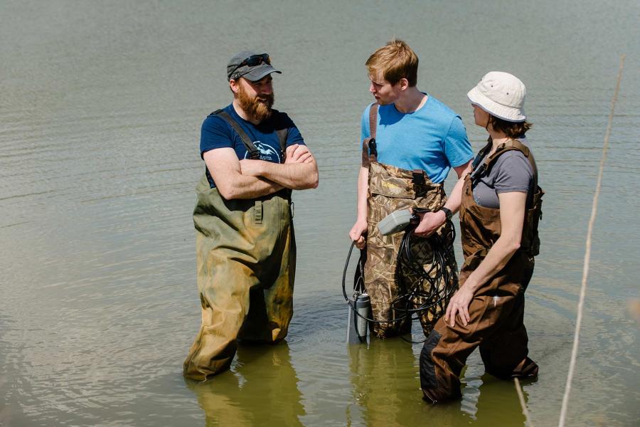 Three researchers wearing hip waders stand talking in knee deep water.