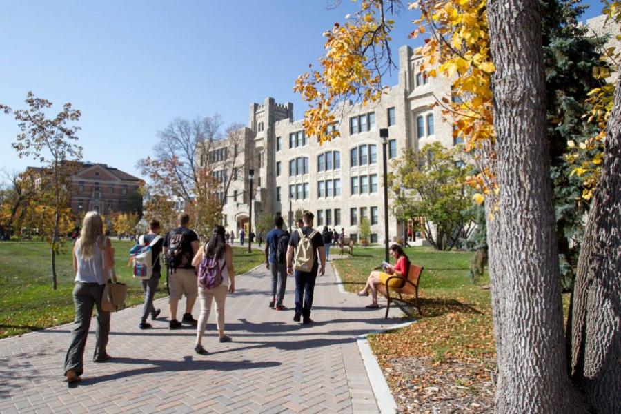 Students walking along a brick path lined with benches and trees in front of a large building.