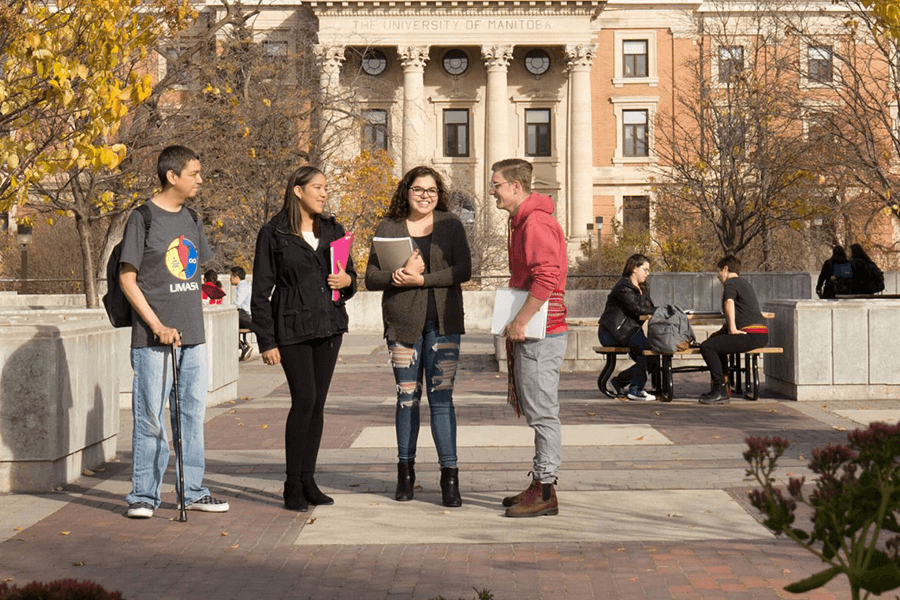 Four students stand together outside of University Centre, several other students are sitting at picnic tables in the background.