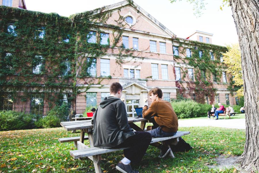 An image of two students sitting on a wooden bench during summer in front of the Engineering building.