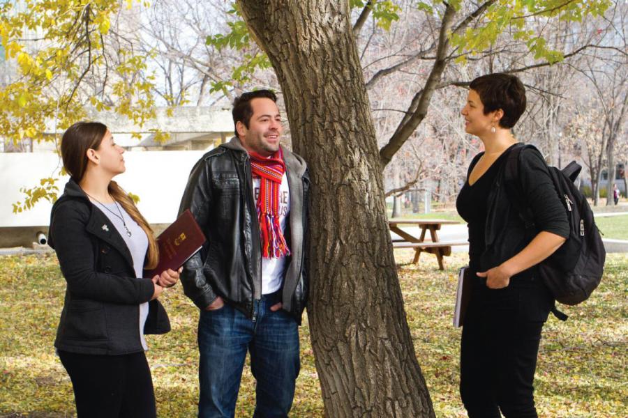 An image of three students standing together outside on a fall day conversing and smiling.