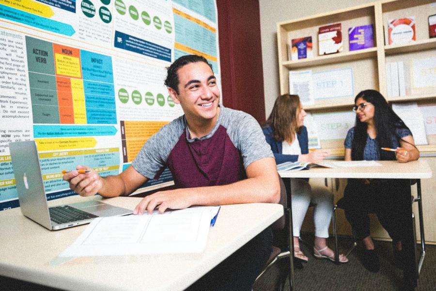 An image of a student sitting at their desk, smiling off to their side, with their laptop and an assignment on their desk.