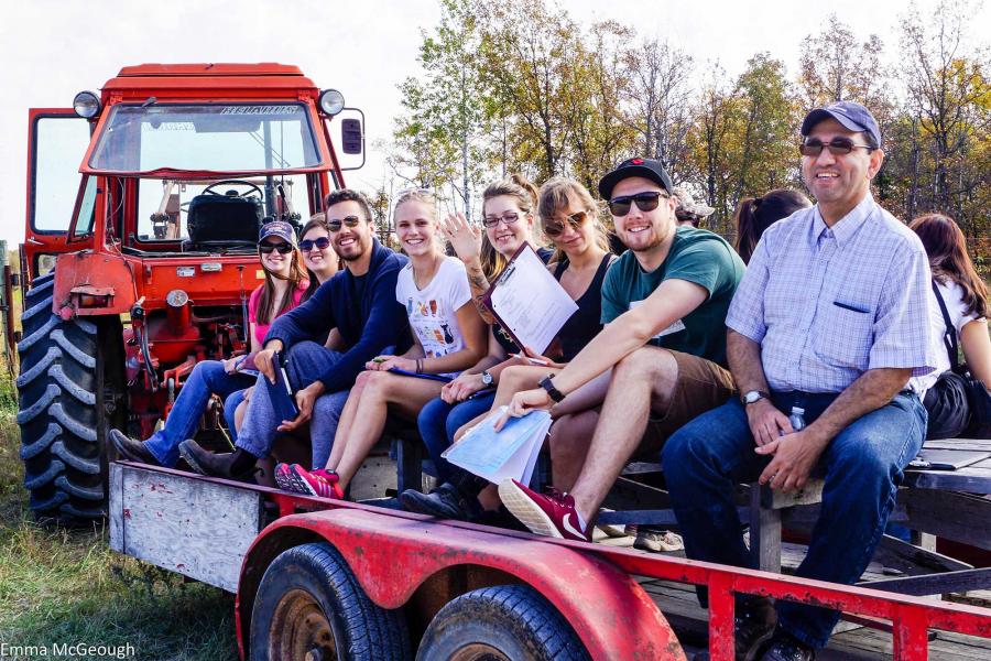 Nine smiling people in summer clothing sit outside on a bench on a flat wooden trailer being pulled by an orange Belarus tractor in a green field.