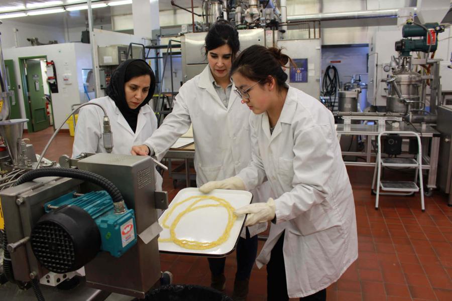 Three people wearing white lab coats stand near a machine in a lab with metal tables. The machine is producing a yellow string of material that one person is catching on a tray.