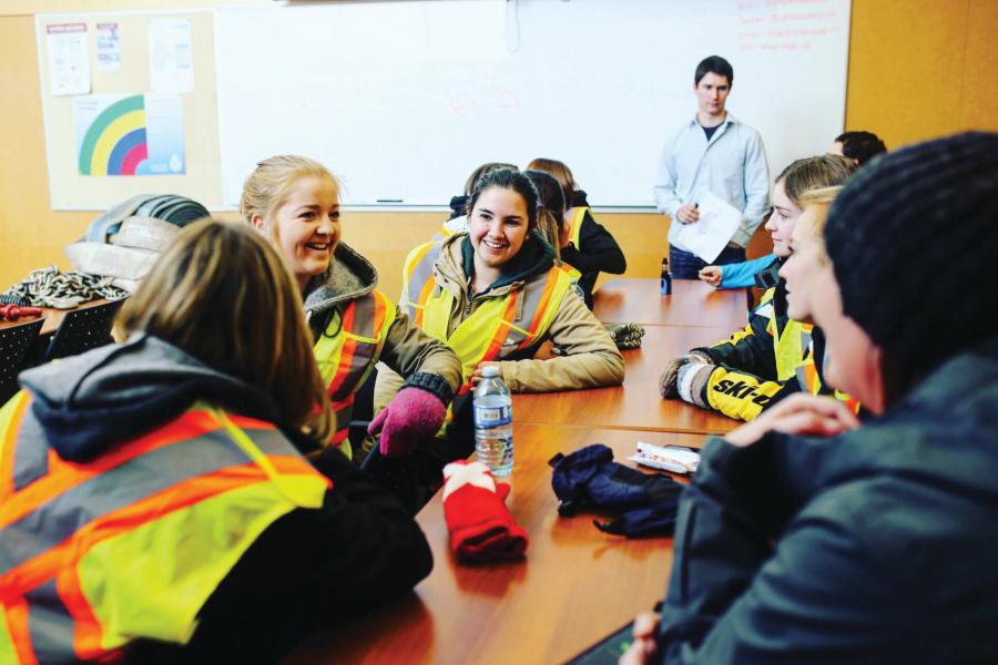 A group of students wearing high visibility vests and outdoor wear sit smiling and talking at a classroom table.