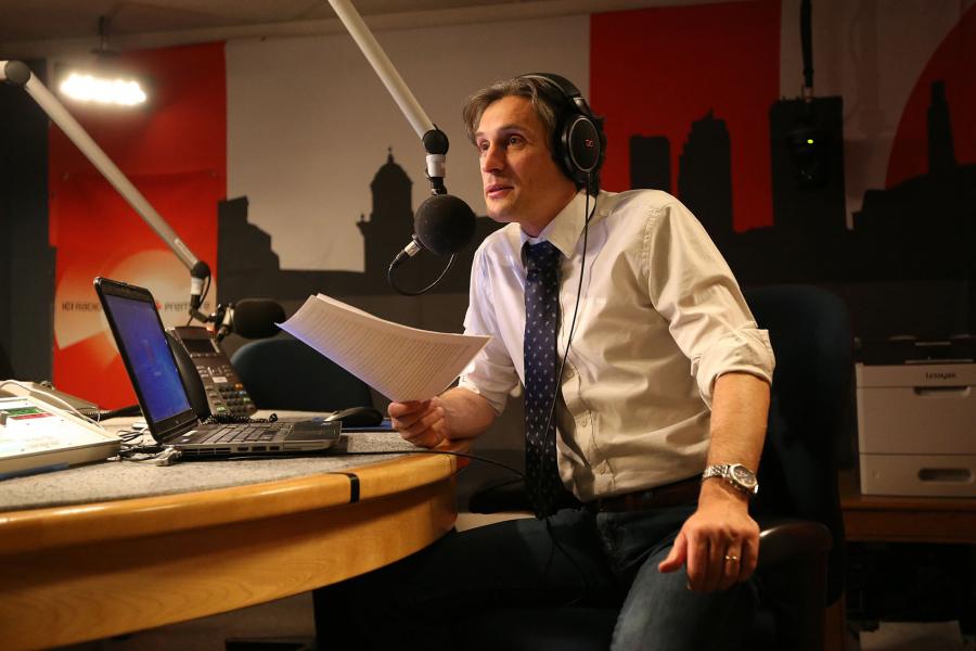 A person wearing a white dress shirt and a blue tie sits at a microphone and laptop reading from a paper script.