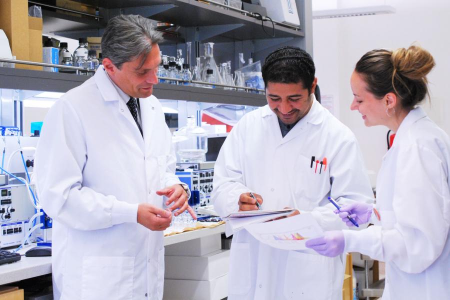 A group of three scientists in white lab coats smile and review graphs in a brightly lit white laboratory.