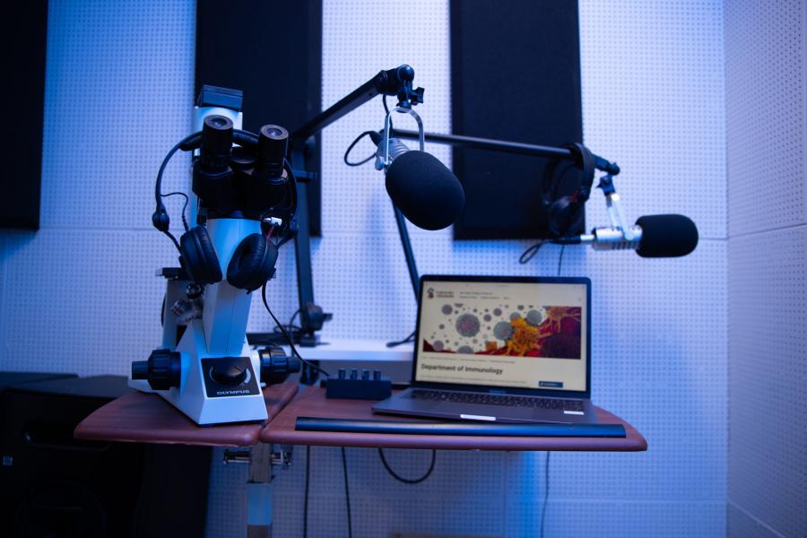 A laptop displays the Department of Immunology website on a table in a recording studio where microphones and headphones hang over a microscope.