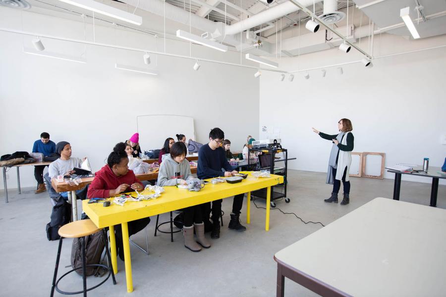 An instructor standing in front of a projector speaks to a class seated at three rows of yellow tables in a white ARTlab room.