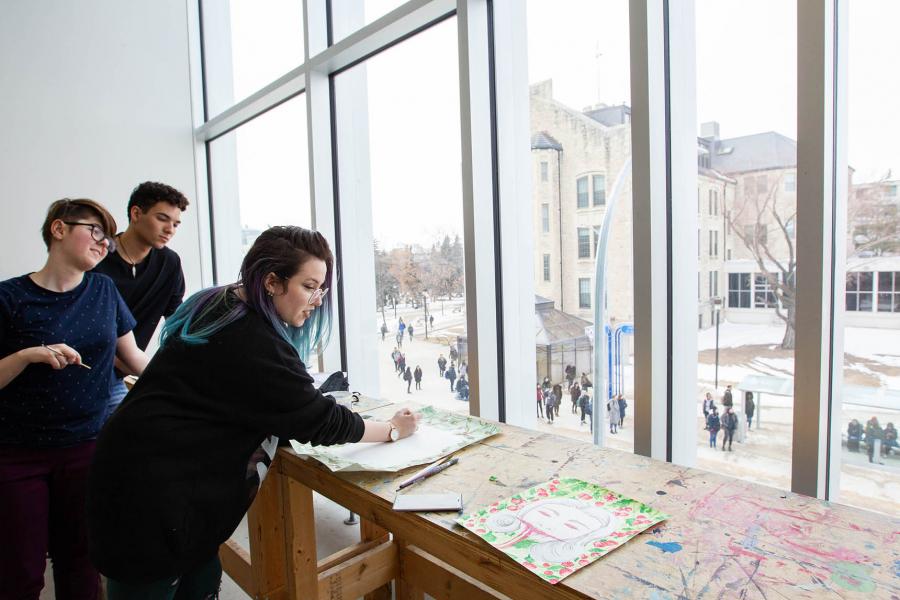 Three students look at drawings on a wooden table that is marked with colourful paint in front of a bright window. The window is facing the bus stop outside of the ARTlab.