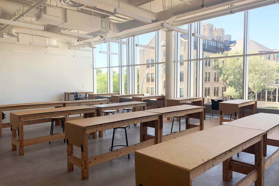 An ARTlab classroom with 10 rows of wooden work tables is bright with sunlight from a large picture window that is facing a brick campus building and green trees.