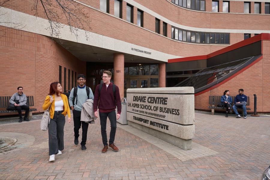 Three students walk together outside of the red brick entryway to the Drake Centre I.H. Asper School of Business building.