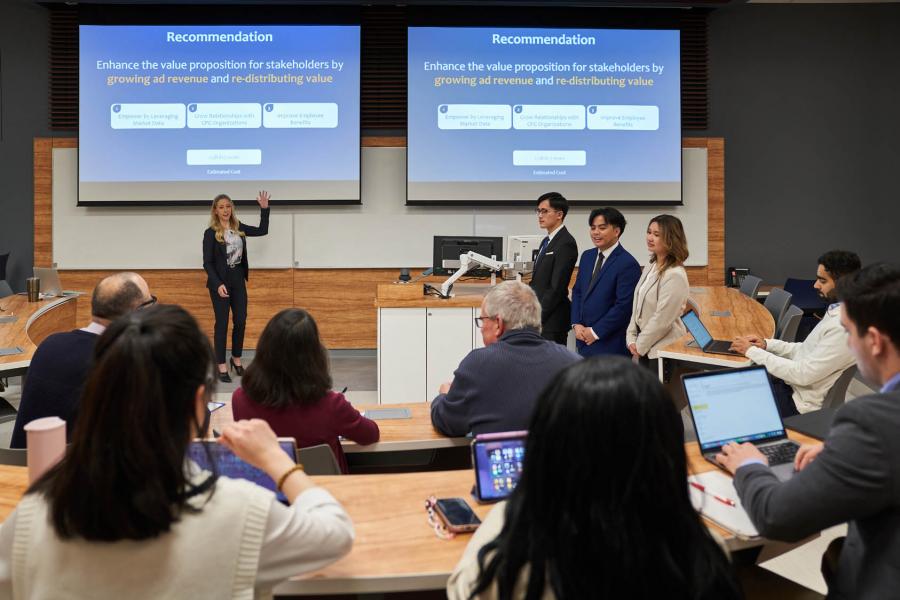 A group of Asper School of Business students stand in front of two presentation screens at the front of a lecture hall audience. The presentation has text that says Recommendation Enhance the value proposition for stakeholders by growing ad revenue and re-distributing value.