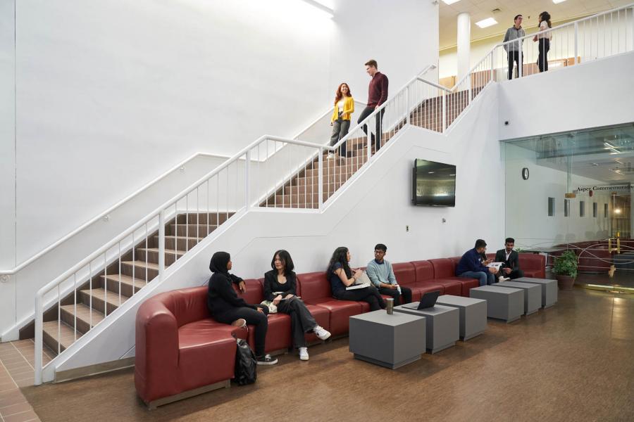 Two students walk down a white staircase heading down to the lobby of the Drake Centre building. The main level has students sitting on red couches in front of gray tables.