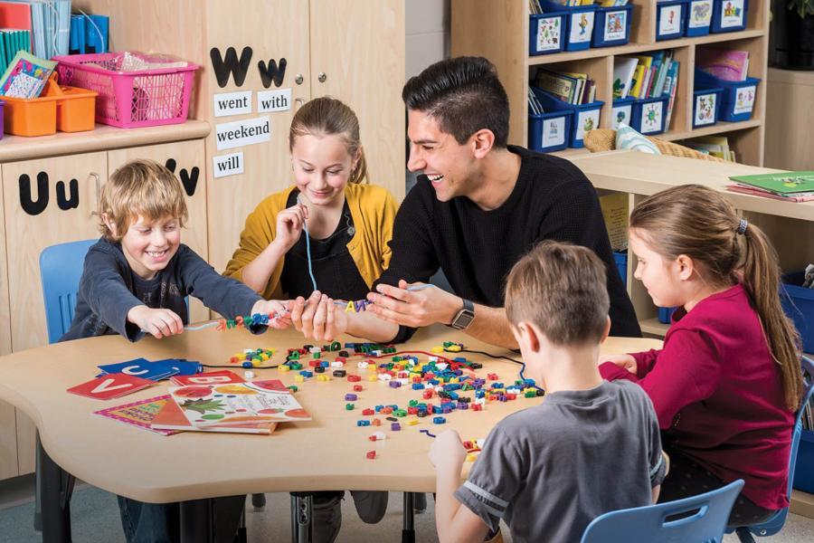 A smiling teacher in a black sweater sits at a small table with four young students that are stringing together letters to spell words on string.