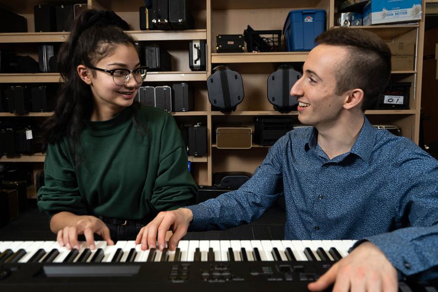 An instructor and a student smile and play a keyboard together in front of a wall of musical instruments in cases.