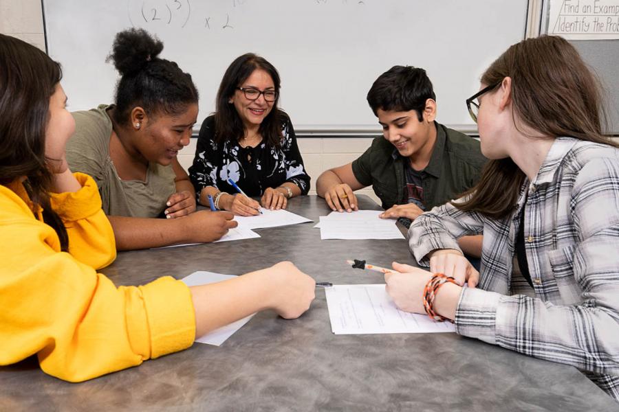 A smiling instructor sits at a gray table with four students who are looking at papers together on the desk.