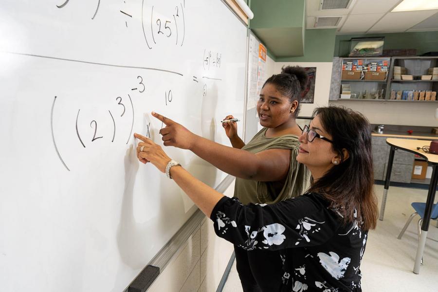 A student who is holding a black marker and a teacher point to an equation on a white dry erase board in an Education Building classroom.