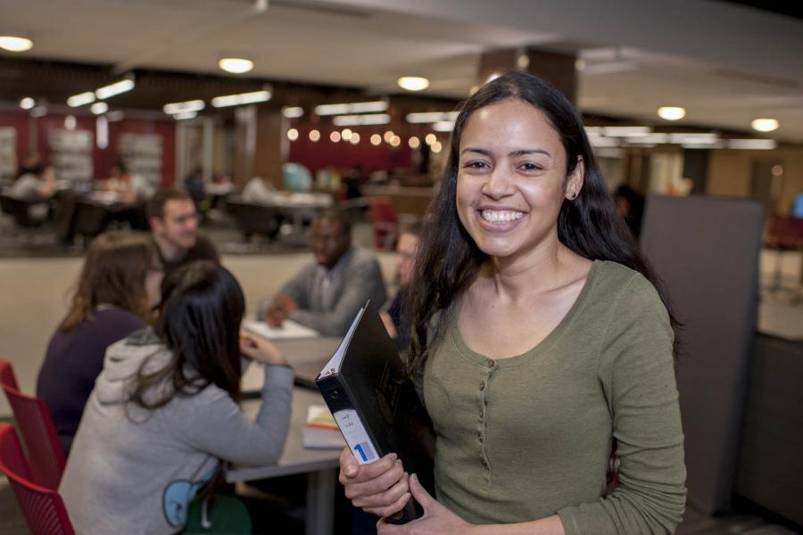 A student wearing a green shirt smiles while holding a black binder in the Elizabeth Dafoe Library.