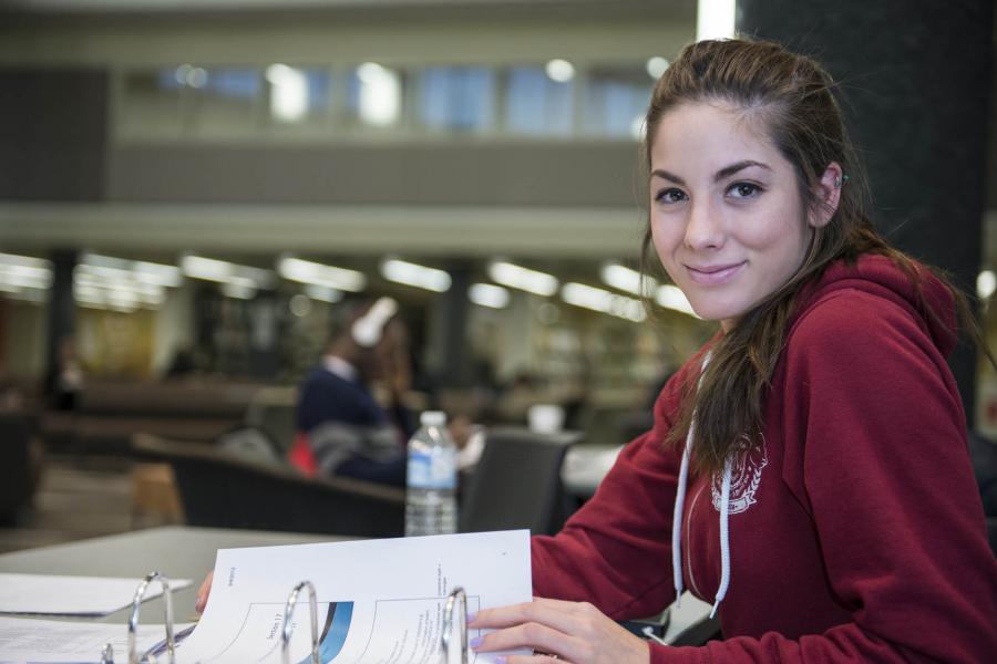 A student wearing a red sweater smiles with the lounge of the Elizabeth Dafoe Library in the background.