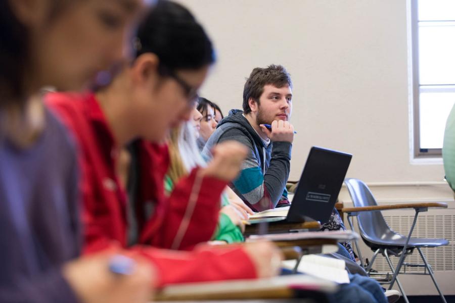 A close up of a student leaning forward attentively on a desk in a row of students in the Fletcher Argue Building.