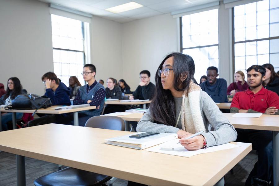 Rows of students at wooden tables that seat two people watch a speaker off screen in a Fletcher Argue Building classroom.