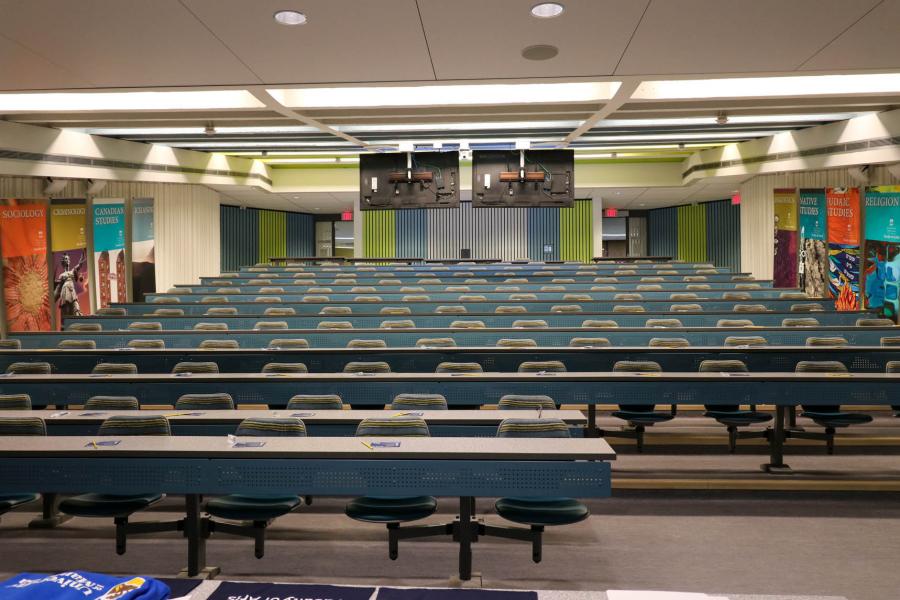 A large, empty lecture hall in the Fletcher Argue Building shows ascending rows of blue seats upwards from the perspective of the speaker.