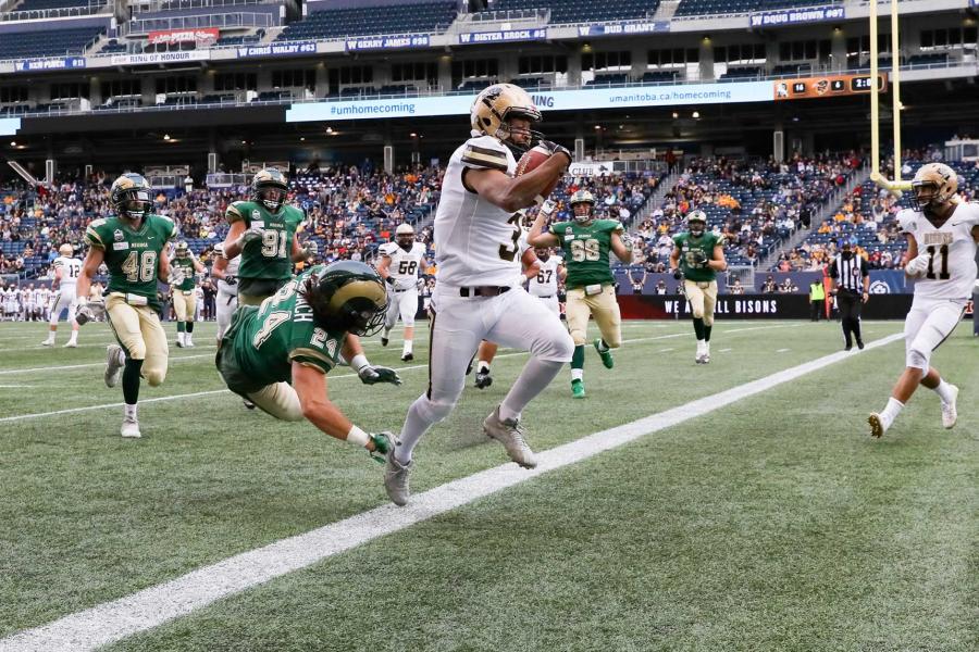 A University of Manitoba Bisons football player runs down the IG Field with a football in his arms past the goalpost while opposing players in green uniforms chase him closely.