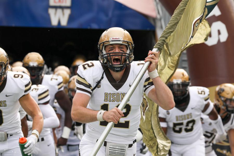 A close up of a University of Manitoba Bisons football player on IG Field. They are wearing a gold helmet, white uniform, and are yelling while holding a gold Bisons flag.