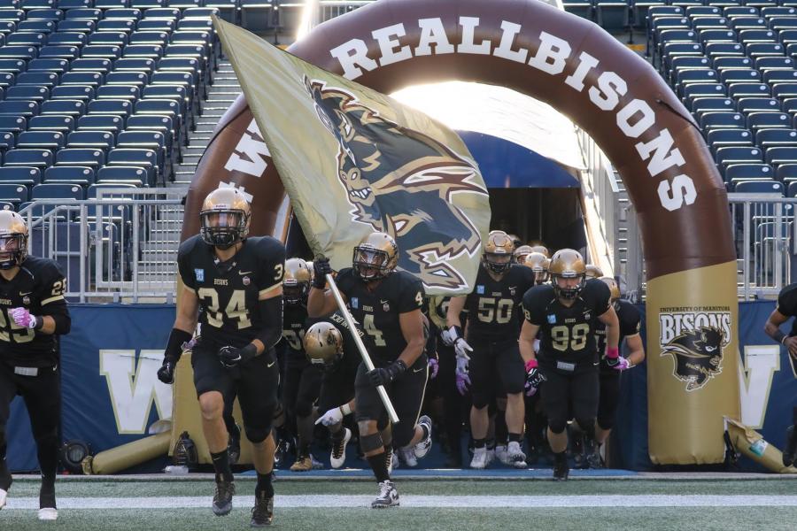 University of Manitoba Bisons football players run onto the IG Field through an inflated archway with the team logo and the words WE ARE ALL BISONS.