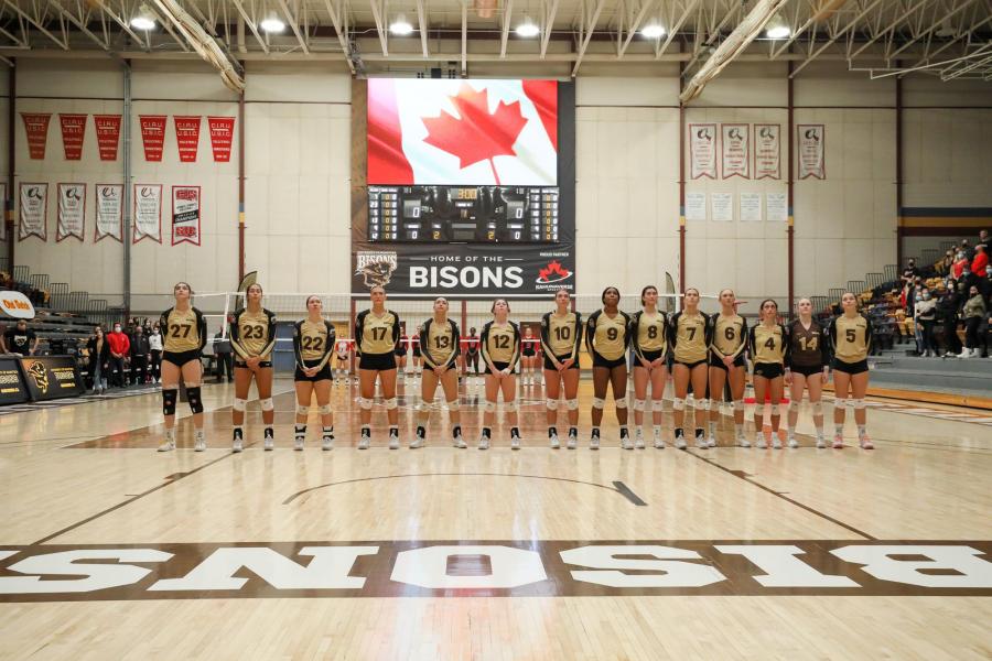 Fourteen members of the UM Bisons women's volleyball team stand in a row on the volleyball court inside of the Investors Group Athletic Centre.