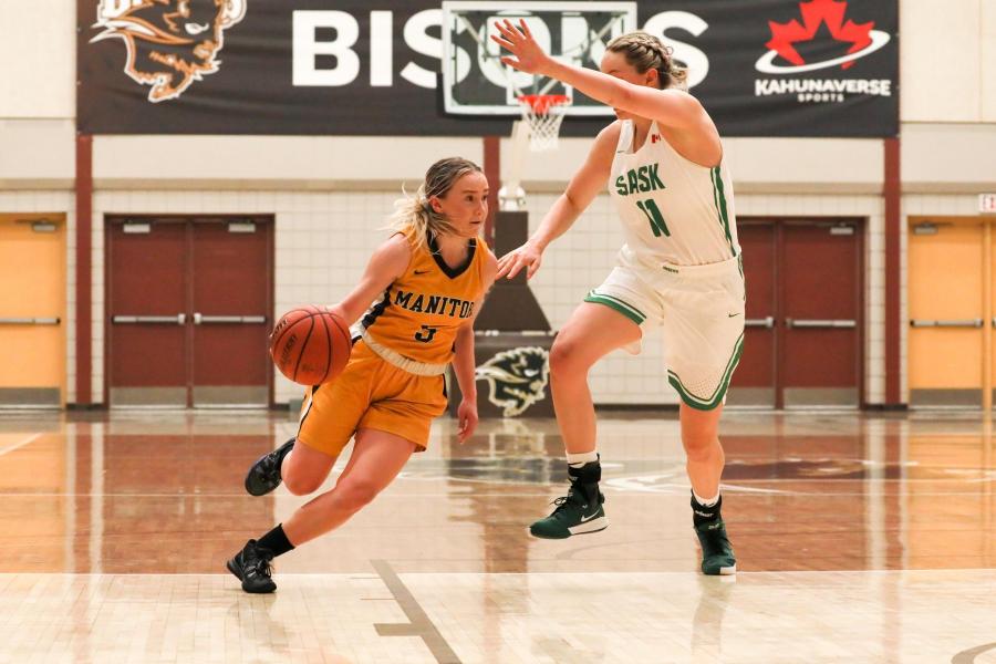 A UM Bisons women's basketball player in a gold jersey dribbles the ball around a Saskatchewan player during a game at the Investors Group Athletic Centre.