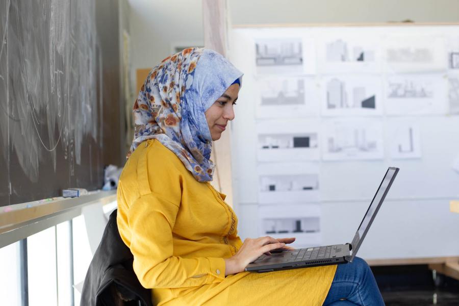 An architecture student wearing a yellow shirt and a colourful blue headscarf types on a laptop in the John A. Russell Building.