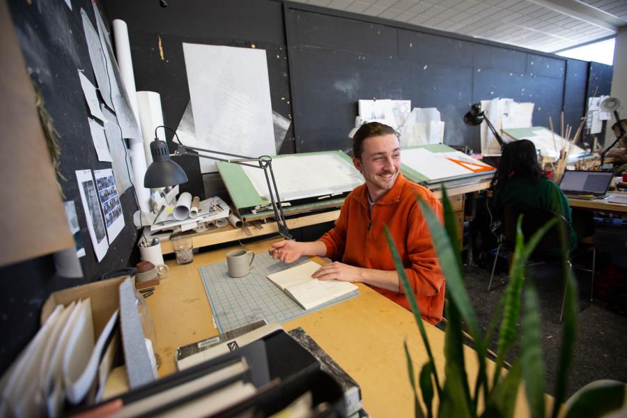 An architecture student wearing an orange shirt smiles while writing in a book in a drafting classroom in the John A. Russell Building.